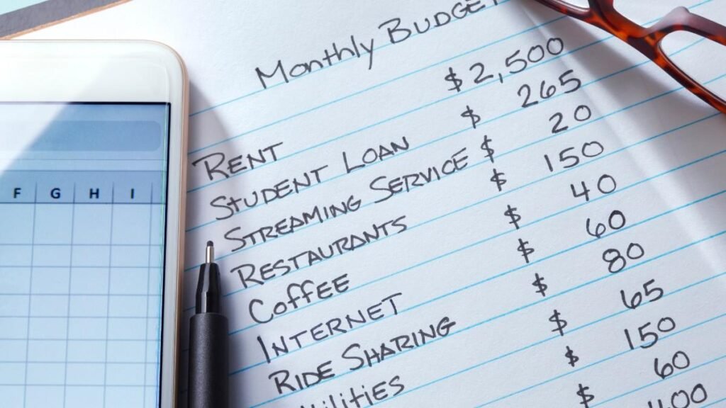 A mobile device and a pen rest on top of a list of monthly expenses written on a sheet of paper. The list reflects a contemporary look at modern day monthly expenses. 